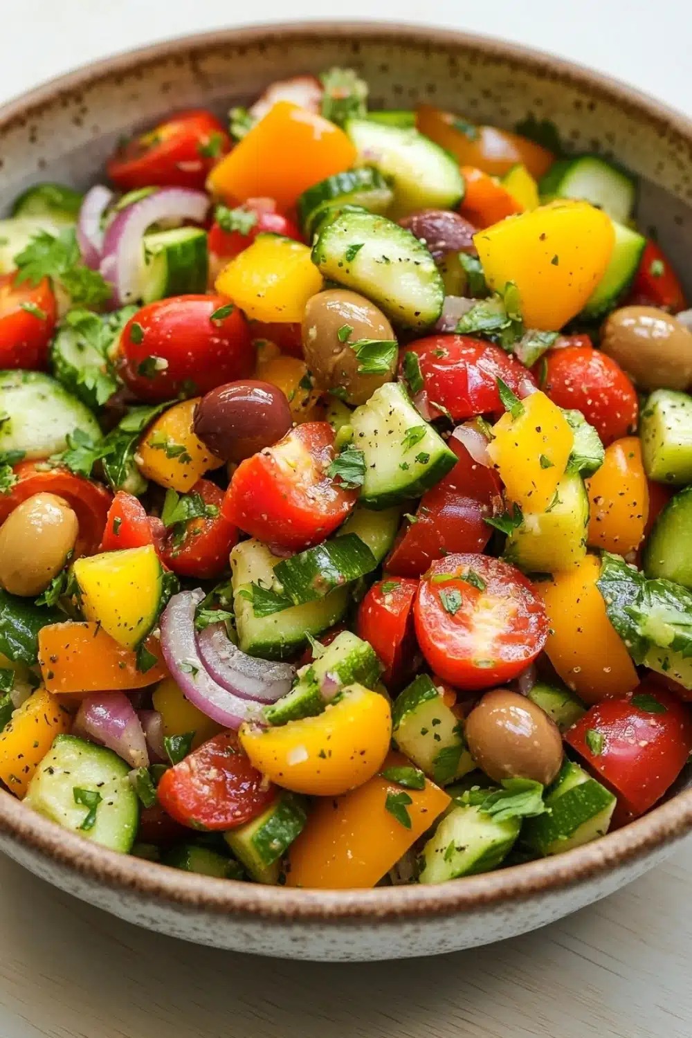 Mediterranean marinated vegetable salad - the image is a close-up of a colorful salad in a bowl. the salad is made up of various types of vegetables, including sliced cucumbers, cherry tomatoes, red onions, black olives, and yellow bell peppers. the vegetables are arranged in a circular pattern, with some overlapping each other. the bowl is made of a light-colored material, possibly ceramic or ceramic, and is sitting on a wooden table. the background is blurred, but it appears to be a kitchen countertop.