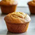 Muffin allo yogurt friggitrice ad aria - the image shows three freshly baked muffins on a baking tray. the muffins are golden brown in color and have a crumbly texture on top. they are arranged in a row on the tray, with one muffin in the foreground and two in the background. the tray is lined with parchment paper and there is a blurred background of a kitchen countertop. the overall mood of the image is warm and inviting.