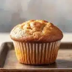 Muffin allo yogurt friggitrice ad aria - the image shows a freshly baked muffin on a wooden tray. the muffin is golden brown in color and has a crumbly texture on top. it appears to be freshly baked and is sitting on a white countertop. the tray is made of metal and has speckled edges. the background is blurred, but it seems to be a kitchen countertop with a gray wall.