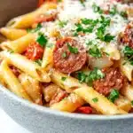 One pot pasta and chorizo - the image is a close-up of a bowl of pasta dish. the pasta appears to be penne, which is a type of penne pasta, and is cooked in a tomato-based sauce. the dish is garnished with chopped parsley and grated parmesan cheese. there are also chunks of sausage and red bell peppers scattered throughout the dish, adding a pop of color and texture. the bowl is made of ceramic and is sitting on a white marble countertop.