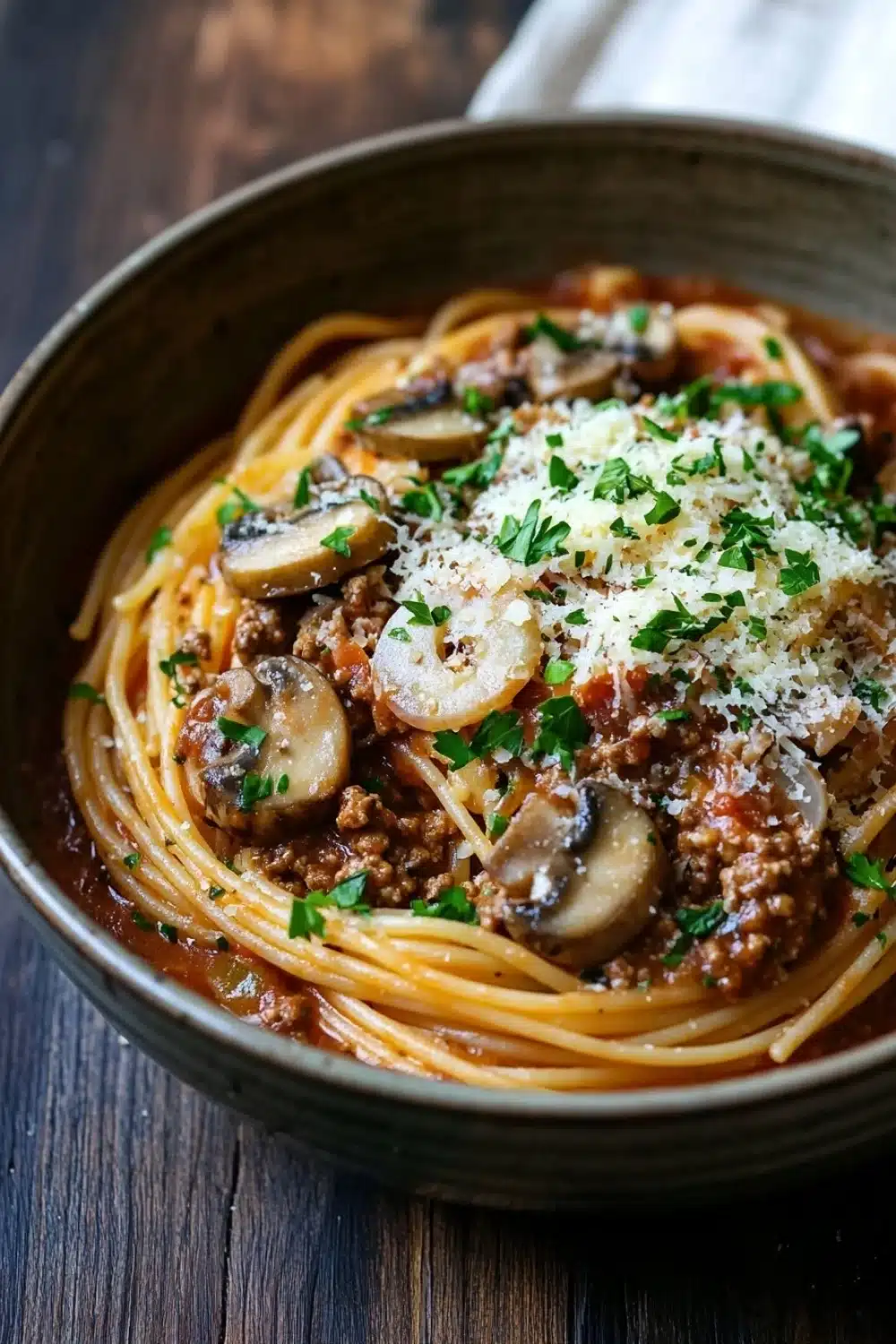 One pot pasta mit hack und pilzen - the image is a close-up of a bowl of spaghetti dish. the spaghetti is cooked al dente and is covered in a rich, red sauce with chunks of meat and mushrooms. the dish is garnished with chopped parsley and grated parmesan cheese. the bowl is sitting on a wooden table with a white napkin in the background. the overall color scheme of the dish is warm and inviting.