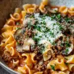 One pot pasta mit hack und pilzen - the image is a close-up of a bowl of pasta dish. the pasta appears to be fusilli, which is a type of spiral-shaped pasta. the dish is made with ground beef, mushrooms, and tomato sauce, and is garnished with chopped herbs and grated parmesan cheese. the bowl is made of ceramic and is sitting on a wooden table. the background is blurred, so the focus is on the dish.