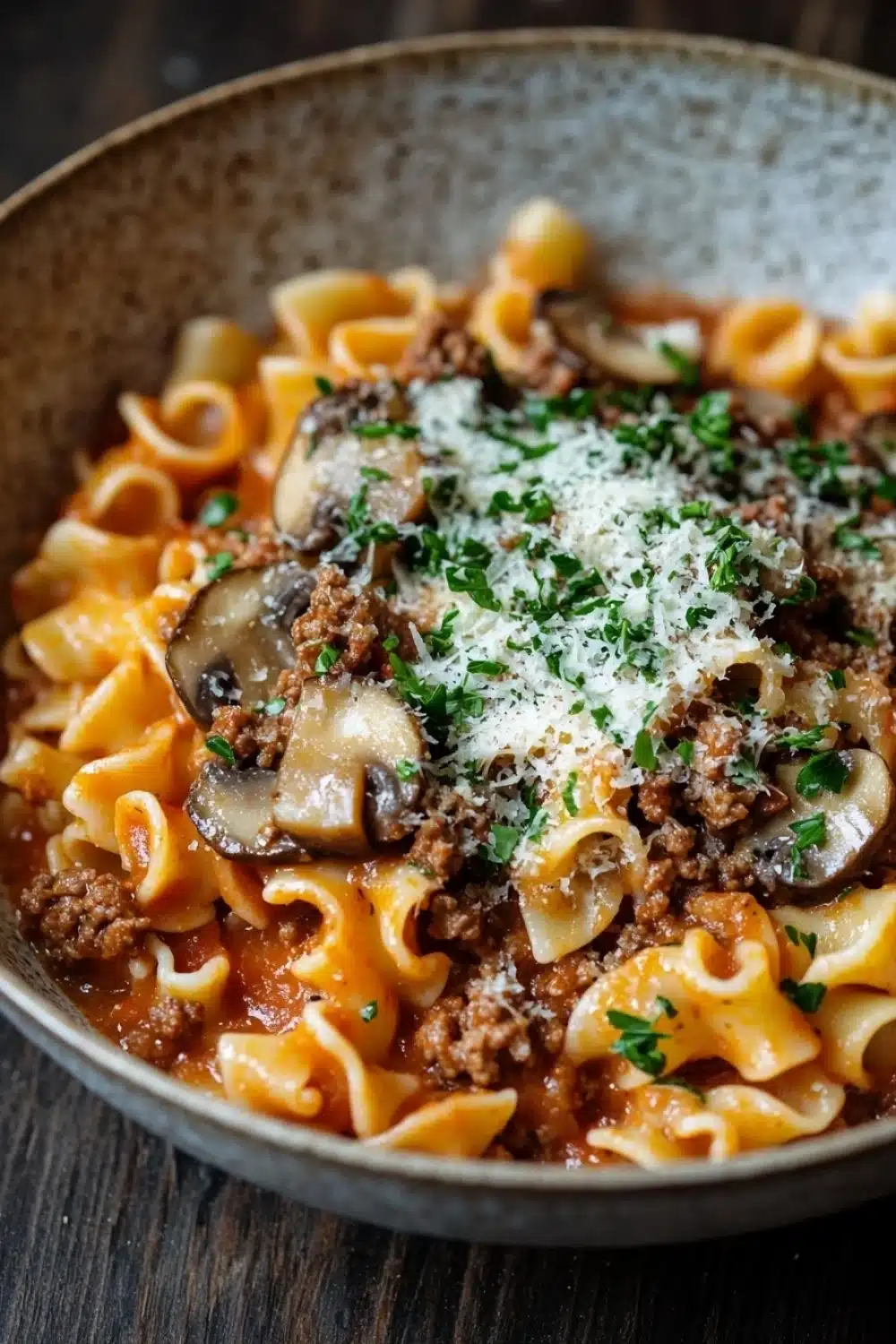 One pot pasta mit hack und pilzen - the image is a close-up of a bowl of pasta dish. the pasta appears to be fusilli, which is a type of spiral-shaped pasta. the dish is made with ground beef, mushrooms, and tomato sauce, and is garnished with chopped herbs and grated parmesan cheese. the bowl is made of ceramic and is sitting on a wooden table. the background is blurred, so the focus is on the dish.