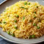 One pot rice - the image shows a plate of yellow rice with peas scattered on top. the rice appears to be cooked and has a fluffy texture. the peas are bright green and look fresh and vibrant. the plate is round and beige in color and is placed on a dark grey textured surface. the background is blurred, making the plate the focal point of the image.