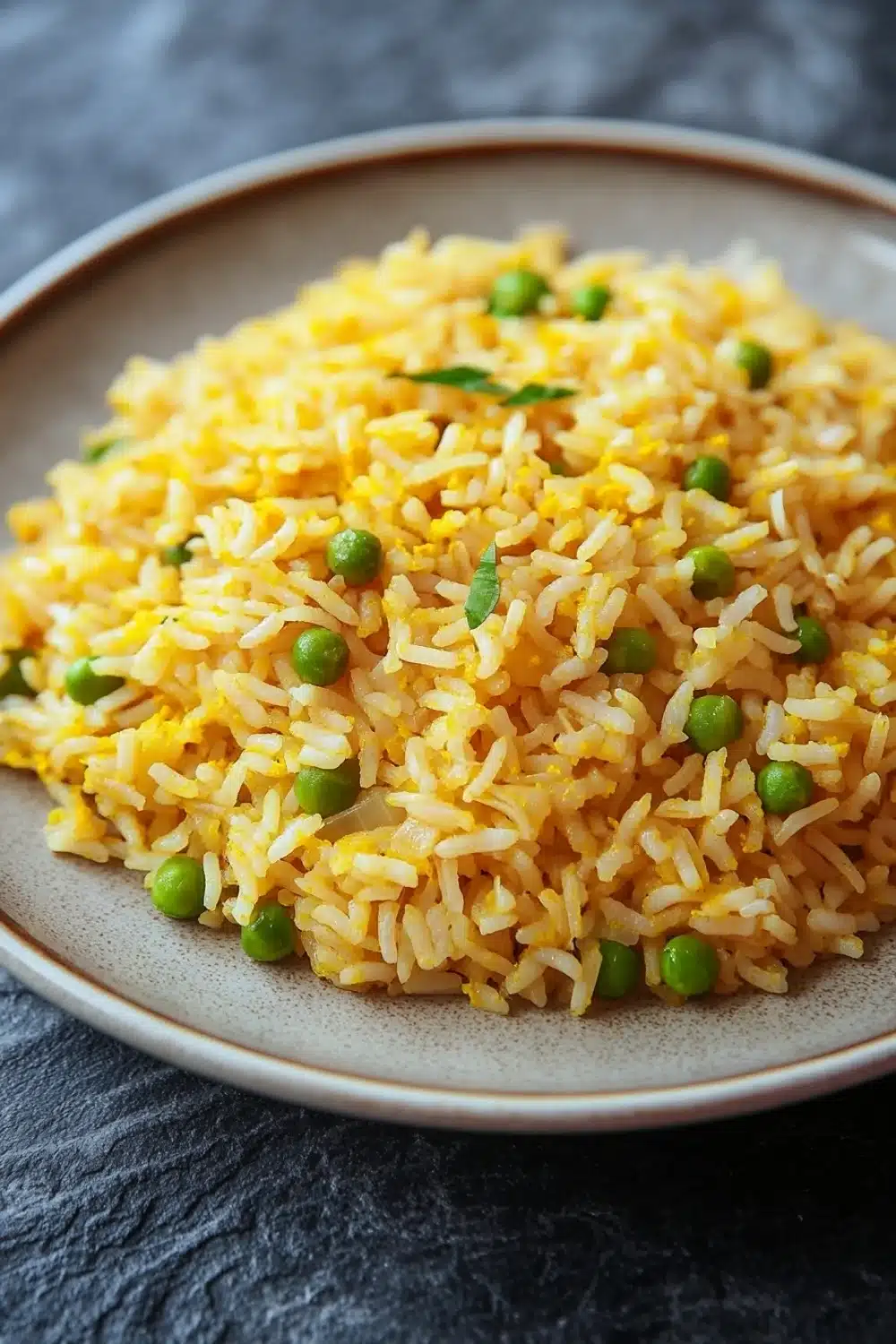 One pot rice - the image shows a plate of yellow rice with peas scattered on top. the rice appears to be cooked and has a fluffy texture. the peas are bright green and look fresh and vibrant. the plate is round and beige in color and is placed on a dark grey textured surface. the background is blurred, making the plate the focal point of the image.