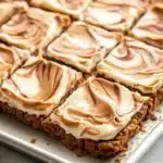 Peanut butter cookie crust dessert - the image shows a rectangular baking tray with a square-shaped dessert on it. the dessert appears to be a graham cracker crust with a layer of peanut butter frosting on top. the frosting is swirled in a swirl pattern and has a light brown color. the tray is lined with parchment paper and is sitting on a white marble countertop.