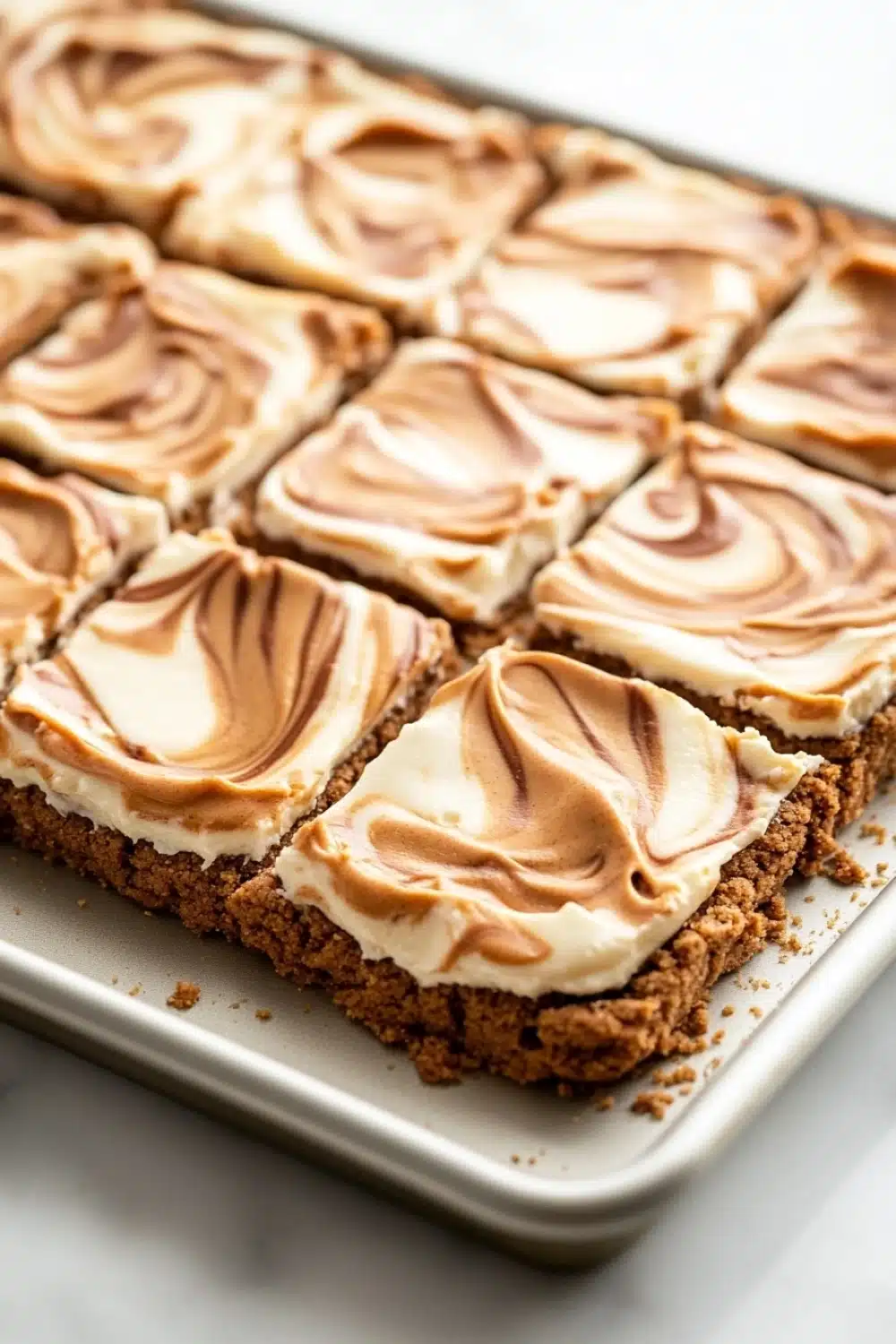 Peanut butter cookie crust dessert - the image shows a rectangular baking tray with a square-shaped dessert on it. the dessert appears to be a graham cracker crust with a layer of peanut butter frosting on top. the frosting is swirled in a swirl pattern and has a light brown color. the tray is lined with parchment paper and is sitting on a white marble countertop.