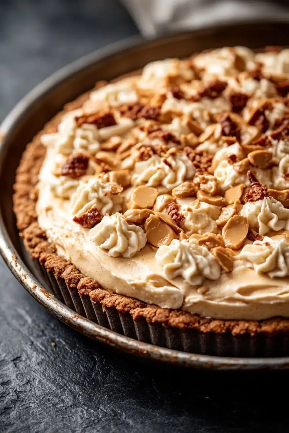 Peanut butter cookie crust dessert - the image is a close-up of a chocolate tart in a round baking dish. the tart has a golden brown crust and is topped with a generous layer of creamy white frosting. the frosting is drizzled with chopped nuts, giving it a crunchy texture. the nuts are scattered on top of the frosting, adding a pop of color to the dish. in the background, there is a white cloth napkin, suggesting that the tart is ready to be served. the overall aesthetic of the image is rustic and appetizing.