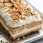 Peanut butter cookie layered dessert - the image is of a dessert on a silver tray. the dessert appears to be a layered dessert with a layer of peanut butter frosting on top. the frosting is white and fluffy, and there are scattered nuts on top of the frosting. the tray is sitting on a white marble countertop.