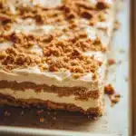 Peanut butter cookie layered dessert - the image shows a rectangular cake on a baking tray. the cake appears to be a layered dessert with a layer of white frosting on top. the frosting is drizzled with a crumbly topping made of chopped nuts. the nuts are arranged in a criss-cross pattern on top of the frosting, giving the cake a crunchy texture. the tray is lined with parchment paper and there are a few crumbs scattered around the tray, indicating that the cake has been freshly baked.