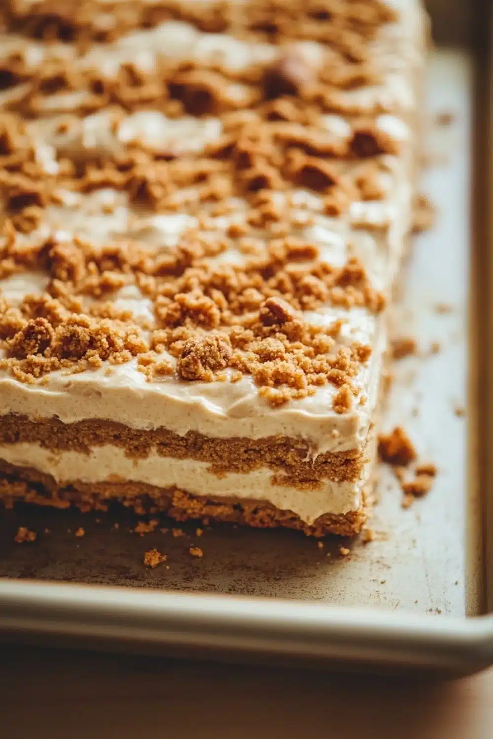 Peanut butter cookie layered dessert - the image shows a rectangular cake on a baking tray. the cake appears to be a layered dessert with a layer of white frosting on top. the frosting is drizzled with a crumbly topping made of chopped nuts. the nuts are arranged in a criss-cross pattern on top of the frosting, giving the cake a crunchy texture. the tray is lined with parchment paper and there are a few crumbs scattered around the tray, indicating that the cake has been freshly baked.