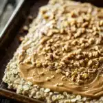 Peanut butter cookie oatmeal - the image shows a rectangular baking tray with a layer of peanut butter on top. the peanut butter is spread evenly over the top of the tray, creating a smooth and creamy texture. the tray is placed on a wooden table, and there are a few scattered nuts around the tray. the background is blurred, but it appears to be a rustic kitchen countertop.