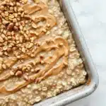 Peanut butter cookie oatmeal - the image shows a rectangular baking dish filled with oatmeal. the oatmeal is light brown in color and has a smooth texture. on top of the oatmeal, there is a generous amount of peanut butter spread in a swirl pattern. there are also a few scattered nuts on top. the dish is sitting on a white marble countertop.