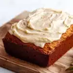Pumpkin bread icing - the image shows a rectangular loaf of bread on a wooden cutting board. the bread appears to be freshly baked and has a golden brown color. on top of the bread, there is a generous layer of white frosting that is spread evenly across the top. the frosting has a smooth texture and looks like it has been freshly baked. there is a sprig of rosemary on the cutting board next to the bread. the background is a white marble countertop.