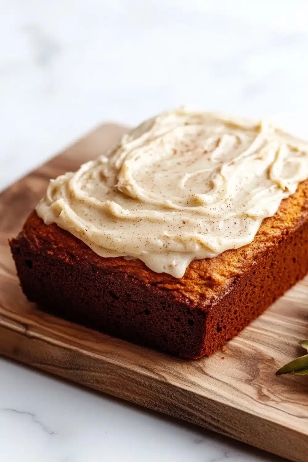 Pumpkin bread icing - the image shows a rectangular loaf of bread on a wooden cutting board. the bread appears to be freshly baked and has a golden brown color. on top of the bread, there is a generous layer of white frosting that is spread evenly across the top. the frosting has a smooth texture and looks like it has been freshly baked. there is a sprig of rosemary on the cutting board next to the bread. the background is a white marble countertop.