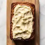 Pumpkin bread icing - the image shows a slice of bread on a wooden cutting board. the bread appears to be freshly baked and has a golden brown crust. on top of the bread, there is a generous amount of creamy white frosting that is spread evenly across the bread. the frosting looks smooth and smooth, and it looks like it has been freshly baked. the cutting board is placed on a white marble countertop.