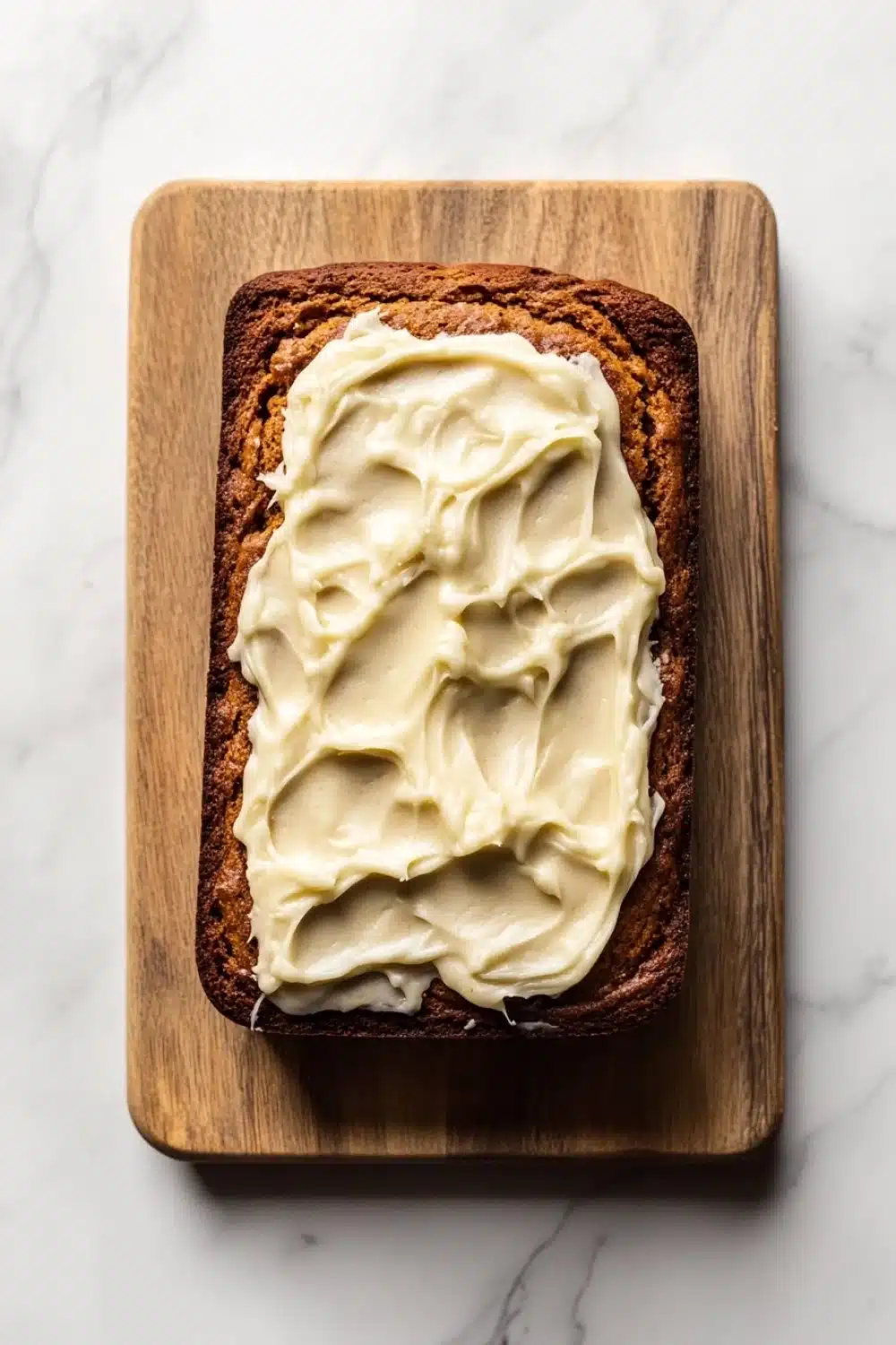 Pumpkin bread icing - the image shows a slice of bread on a wooden cutting board. the bread appears to be freshly baked and has a golden brown crust. on top of the bread, there is a generous amount of creamy white frosting that is spread evenly across the bread. the frosting looks smooth and smooth, and it looks like it has been freshly baked. the cutting board is placed on a white marble countertop.
