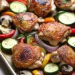 Sheet pan hibachi chicken and vegetables - the image shows a baking tray filled with grilled chicken and vegetables. the chicken is golden brown and appears to be seasoned with herbs and spices. the vegetables include sliced zucchini, red bell peppers, and mushrooms. the tray is lined with parchment paper and is sitting on a wooden table. the background is blurred, but it seems to be a kitchen countertop.