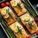 Sheet pan lemon brown butter salmon - the image shows four pieces of salmon fillets on a baking tray. the salmon is cooked to a golden brown color and is garnished with lemon wedges and fresh herbs. the tray is lined with asparagus spears and cherry tomatoes, and there is a sprig of thyme on the side. the background is a dark grey countertop with a white cloth napkin and a black and white striped napkin on the right side.