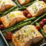 Sheet pan lemon brown butter salmon - the image shows a baking tray with six pieces of salmon and asparagus. the salmon is cooked to a golden brown color and appears to be seasoned with herbs and spices. the asparaglips are bright green and are arranged in a single layer on the tray. there are also small red cherry tomatoes scattered around the tray, adding a pop of color to the dish. the tray is sitting on a dark grey countertop.