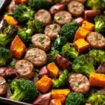 Sheet pan sausage sweet potato broccoli - the image shows a tray of cooked vegetables on a wooden table. the vegetables appear to be broccoli, sweet potatoes, and sausage. the sausage is cut into small cubes and is arranged in a circular pattern on the tray. the broccoli is bright green and the sweet potatoes are orange. the carrots are cut into thin slices and are scattered throughout the tray, with some of them overlapping each other. the tray is lined with parchment paper and there is a sprinkle of herbs on top. the background is blurred, but it appears to be a kitchen countertop.