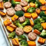 Sheet pan sausage sweet potato broccoli - the image shows a baking tray lined with parchment paper. on the tray, there are several pieces of roasted vegetables, including sweet potatoes, broccoli, and sausage. the vegetables are arranged in a single layer on the tray and appear to be seasoned with herbs and spices. the tray is sitting on a white countertop.
