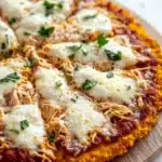 Shredded chicken keto pizza crust - the image is of a freshly baked pizza on a wooden cutting board. the pizza has a golden brown crust and is topped with shredded chicken and melted cheese. the chicken is golden brown and appears to be seasoned with herbs and spices. the cheese is melted and bubbly, and there are small chunks of tomato sauce scattered throughout the pizza. the cutting board is placed on a white wooden table with a white cloth napkin in the background.
