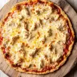 Shredded chicken keto pizza crust - the image is of a round pizza on a wooden cutting board. the pizza has a golden brown crust and is topped with melted cheese and chunks of chicken. the cheese appears to be melted and bubbly, and the chicken is visible on top. the cutting board is placed on a white wooden table. the background is blurred, making the pizza the focal point of the image.
