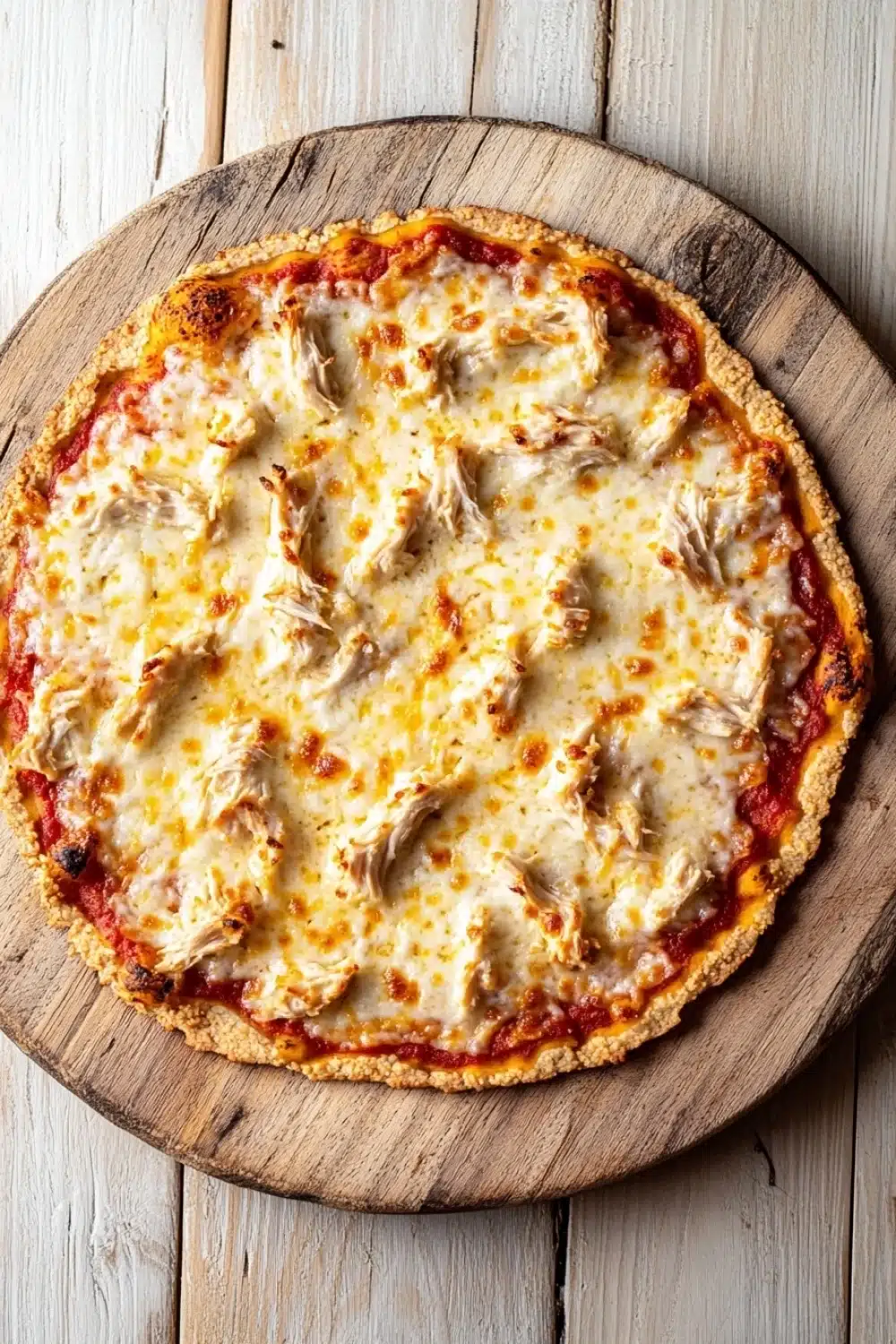 Shredded chicken keto pizza crust - the image is of a round pizza on a wooden cutting board. the pizza has a golden brown crust and is topped with melted cheese and chunks of chicken. the cheese appears to be melted and bubbly, and the chicken is visible on top. the cutting board is placed on a white wooden table. the background is blurred, making the pizza the focal point of the image.