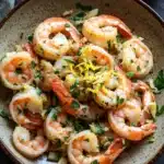 Skillet shrimp scampi - the image is a close-up of a bowl of cooked shrimp. the shrimp are orange in color and appear to be seasoned with herbs and spices. the bowl is made of a light-colored ceramic with a speckled pattern. there is a slice of lemon on the side of the bowl and a gray cloth napkin on the left side. the background is a dark grey countertop.