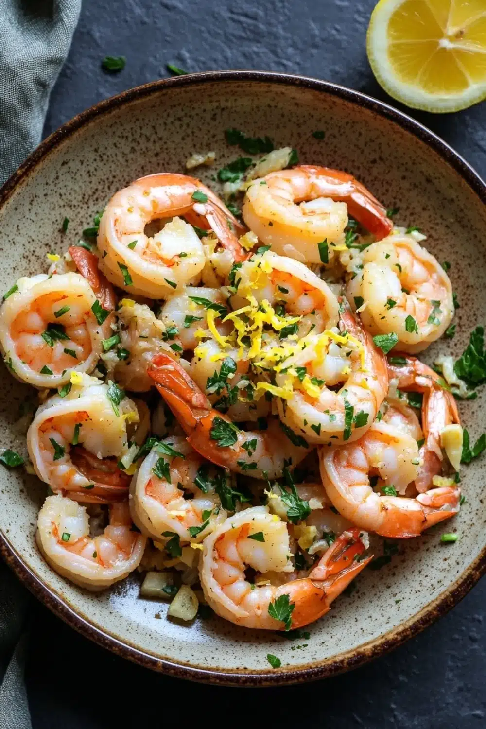 Skillet shrimp scampi - the image is a close-up of a bowl of cooked shrimp. the shrimp are orange in color and appear to be seasoned with herbs and spices. the bowl is made of a light-colored ceramic with a speckled pattern. there is a slice of lemon on the side of the bowl and a gray cloth napkin on the left side. the background is a dark grey countertop.