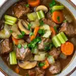 Slow cooker beef mushroom - the image is a close-up of a bowl of beef stew. the stew is made with chunks of beef, carrots, celery, onions, and mushrooms. the beef is cooked in a rich, dark brown broth and is garnished with fresh parsley. the vegetables are cut into small pieces and are arranged in a circular pattern around the beef. the bowl is made of ceramic and has a brown rim. the background is a white marble countertop.