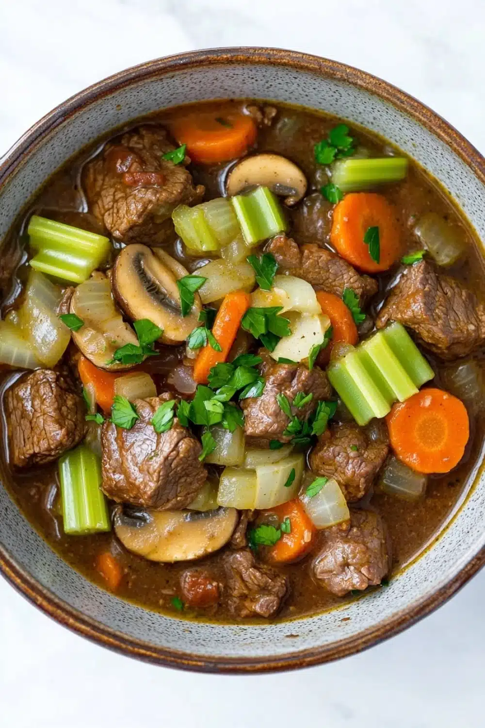 Slow cooker beef mushroom - the image is a close-up of a bowl of beef stew. the stew is made with chunks of beef, carrots, celery, onions, and mushrooms. the beef is cooked in a rich, dark brown broth and is garnished with fresh parsley. the vegetables are cut into small pieces and are arranged in a circular pattern around the beef. the bowl is made of ceramic and has a brown rim. the background is a white marble countertop.