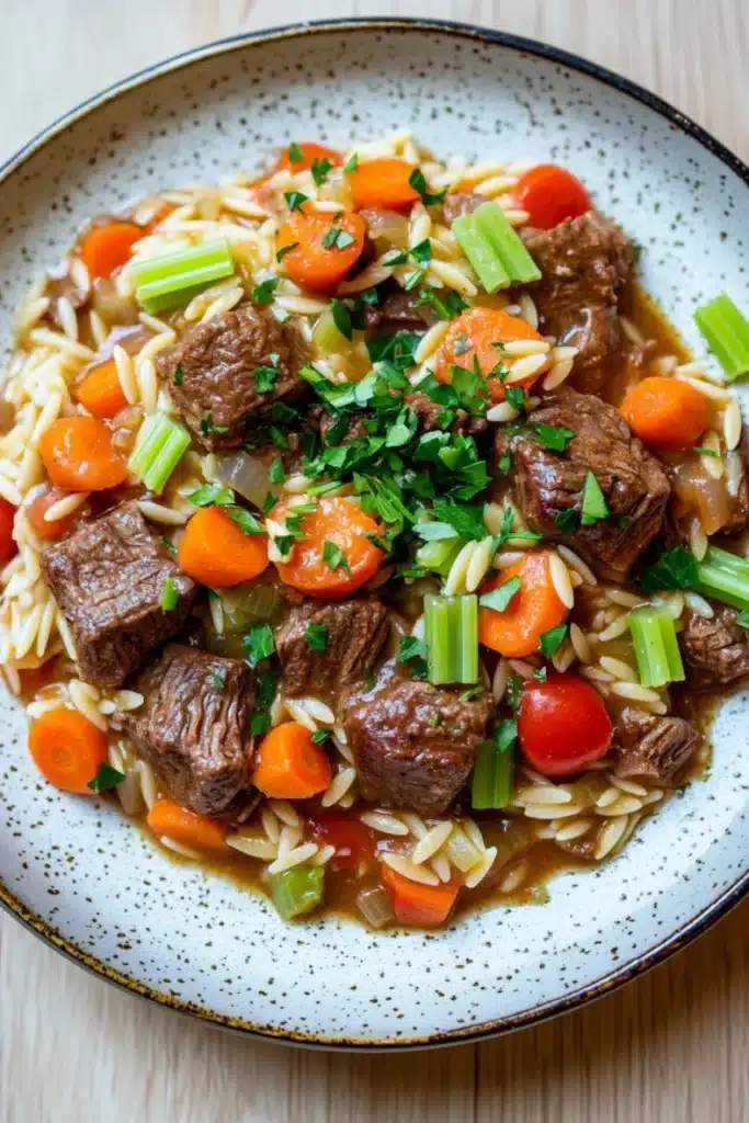Slow cooker beef orzo - the image is a close-up of a bowl of beef and vegetable stir-fry. the bowl is white with a speckled pattern and has a blue rim. the beef is cooked medium-rare and appears to be beef tenderloin, with chunks of carrots, celery, and onions mixed in. the vegetables are bright orange and look fresh and vibrant. the dish is served on a bed of fluffy white noodles. the background is a wooden table.