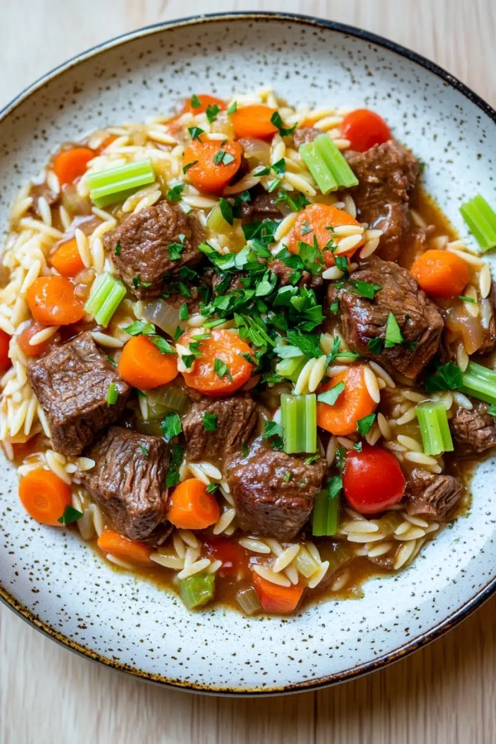 Slow cooker beef orzo - the image is a close-up of a bowl of beef and vegetable stir-fry. the bowl is white with a speckled pattern and has a blue rim. the beef is cooked medium-rare and appears to be beef tenderloin, with chunks of carrots, celery, and onions mixed in. the vegetables are bright orange and look fresh and vibrant. the dish is served on a bed of fluffy white noodles. the background is a wooden table.