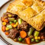 Slow cooker beef pot pie - the image shows a plate of beef stew with a golden brown crust on top. the beef stew is made with chunks of beef, carrots, peas, corn, and other vegetables, all cooked in a rich, dark brown sauce. the dish is served on a white plate with a brown rim. the background is blurred, but it appears to be a kitchen countertop. the overall color scheme of the dish is warm and inviting.
