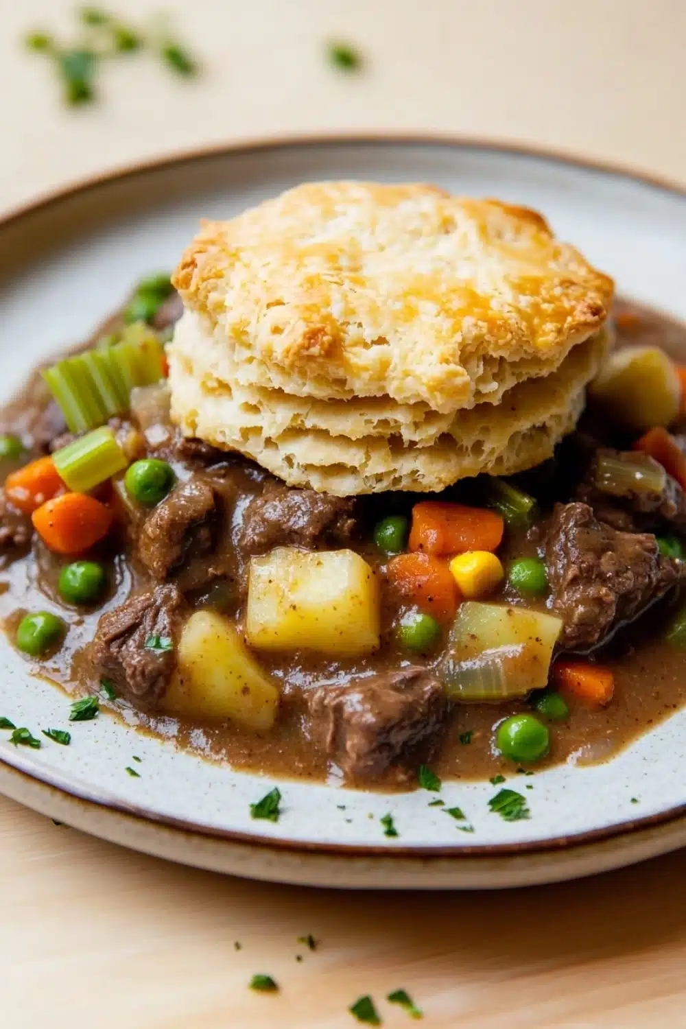 Slow cooker beef pot pie - the image shows a plate of beef stew with a biscuit on top. the stew is made with chunks of beef, potatoes, carrots, and peas, and is garnished with chopped parsley. the plate is white with a blue rim and is sitting on a wooden table. the biscuits are golden brown and appear to be freshly baked. the background is blurred, but it appears to be a kitchen countertop.