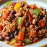 Slow cooker turkey quinoa chili - the image is a close-up of a plate of food. the plate is white with a gold rim and is placed on a wooden table. the food appears to be a dish made with ground beef, black beans, red and green bell peppers, and other vegetables. the beef is cooked to a golden brown color and is topped with a sprinkle of quinoa. the vegetables are cut into small cubes and are scattered throughout the dish. the dish looks appetizing and ready to eat.
