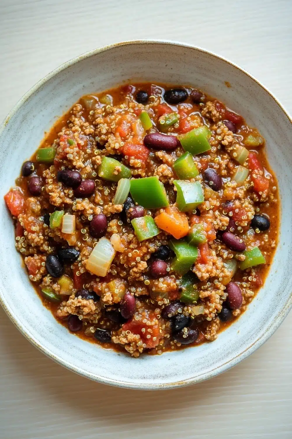 Slow cooker turkey quinoa chili - the image is a close-up of a bowl of chili. the bowl is made of ceramic and has a light blue rim. the chili is made up of quinoa, black beans, red kidney beans, diced carrots, and green bell peppers. the quinoa is a reddish-brown color and appears to be cooked until it is soft and fluffy. there are also small pieces of diced onions scattered throughout the chili, adding a pop of color to the dish. the dish is sitting on a white wooden table.