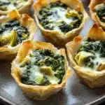 Spinach artichoke dip filo cups - the image shows a plate of small, golden-brown wonton cups filled with a mixture of spinach and artichokes. the wontons are arranged in a circular pattern on the plate, with some overlapping each other. the spinach is a vibrant green color and appears to be cooked until it is soft and fluffy. the cheese is melted and bubbly, and the wontons have a golden brown crust on top. the plate is sitting on a dark, textured surface.