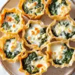 Spinach artichoke dip filo cups - the image shows a plate of mini quiches on a white marble countertop. the quiche shells are golden brown and have a flaky crust. they are filled with a mixture of spinach, cheese, and other ingredients. the spinach is a vibrant green color and appears to be cooked until it is soft and fluffy. the cheese is melted and bubbly, and there are small pieces of orange-colored cheese scattered throughout the dish. the plate is garnished with fresh parsley leaves.