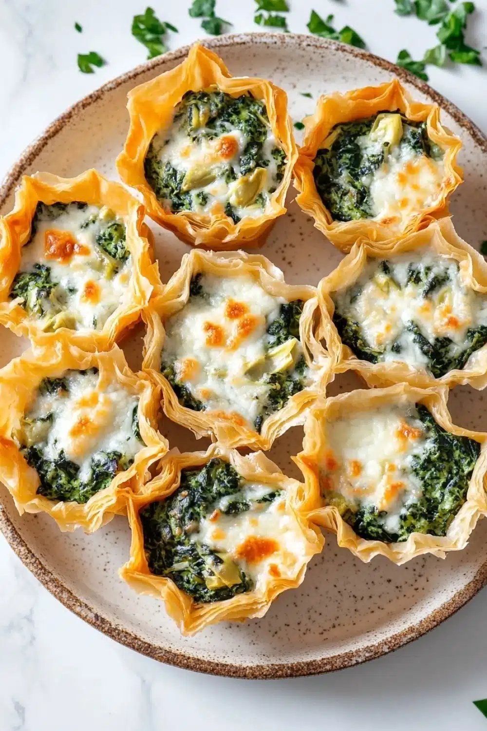 Spinach artichoke dip filo cups - the image shows a plate of mini quiches on a white marble countertop. the quiche shells are golden brown and have a flaky crust. they are filled with a mixture of spinach, cheese, and other ingredients. the spinach is a vibrant green color and appears to be cooked until it is soft and fluffy. the cheese is melted and bubbly, and there are small pieces of orange-colored cheese scattered throughout the dish. the plate is garnished with fresh parsley leaves.