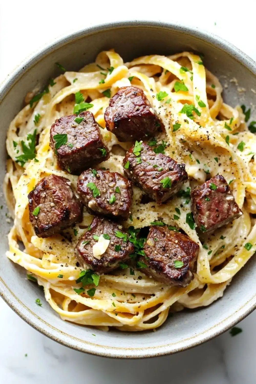 Steak bites with garlic parmesan pasta - the image is a close-up of a bowl of pasta dish. the pasta appears to be fettuccine alfredo, which is a type of italian pasta. the dish is garnished with chopped parsley and a sprinkle of parmesan cheese. the meat is cooked medium-rare and has a dark brown color. the bowl is made of ceramic and is sitting on a white marble countertop.