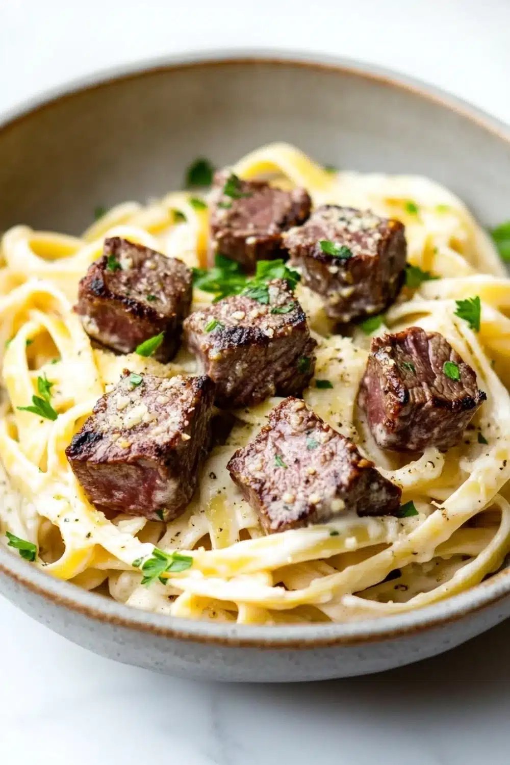 Steak bites with garlic parmesan pasta - the image is a close-up of a bowl of pasta dish. the pasta appears to be fettuccine alfredo, which is a type of italian pasta. the dish is garnished with chopped parsley and a sprinkle of herbs. the meat is cooked medium-rare and has a dark brown color. the bowl is made of ceramic and is sitting on a white marble countertop. the overall presentation of the dish is elegant and appetizing.