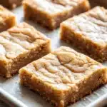 Sugar cookie bar - the image shows a baking tray with six square-shaped bars on it. the bars are golden brown in color and have a crumbly texture on top. they appear to be freshly baked and are arranged in a neat row on the tray. the tray is lined with parchment paper and is placed on a wooden table. the background is blurred, but it appears to be a kitchen countertop.