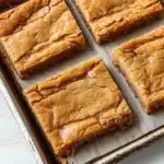 Sugar cookie bar - the image shows a baking tray lined with square-shaped brownies. the brownies have a crumbly texture and appear to be freshly baked. they are arranged in a single layer on the tray, with some overlapping each other. the tray is placed on a white wooden surface, and the background is blurred, making the brownies the focal point of the image.
