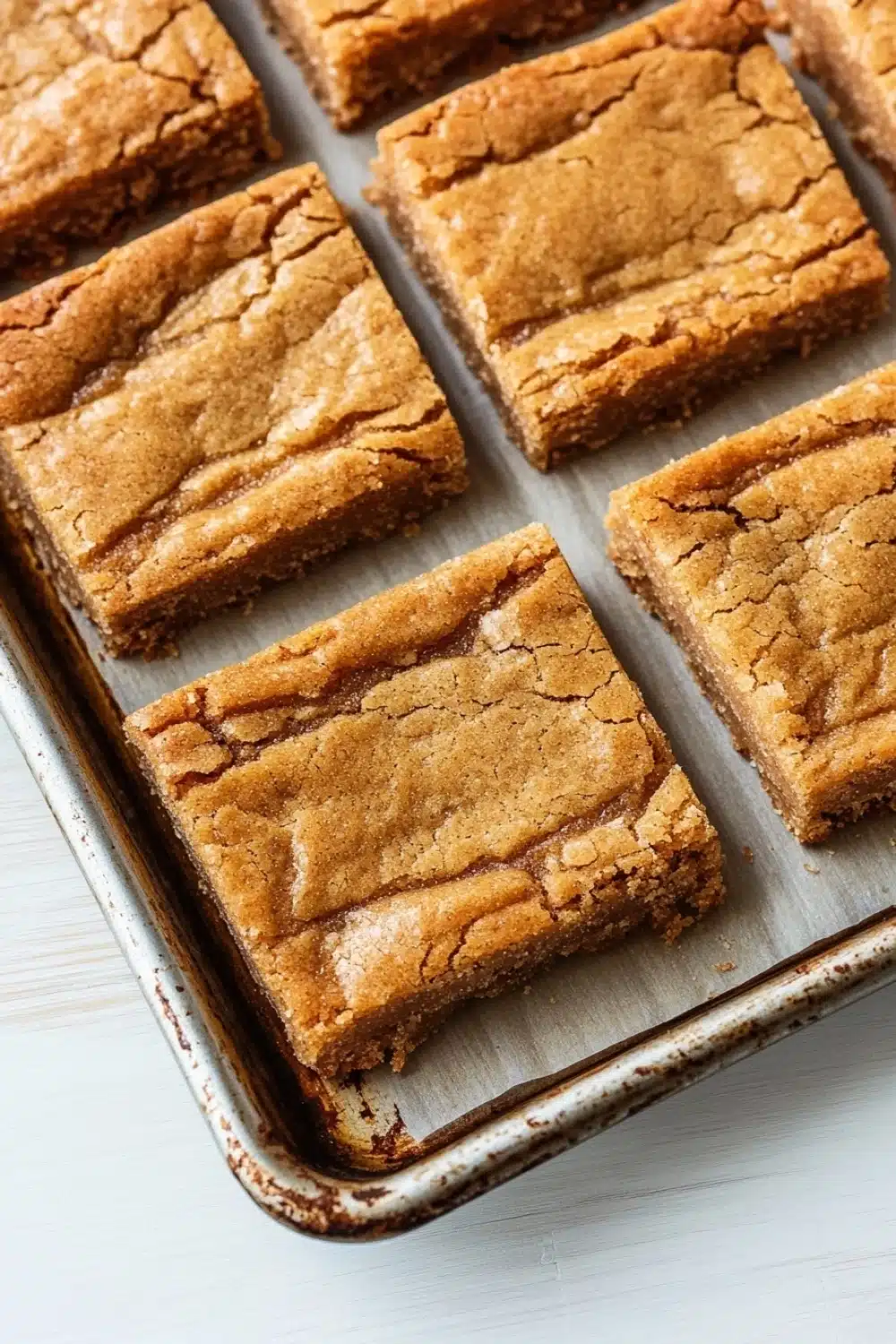 Sugar cookie bar - the image shows a baking tray lined with square-shaped brownies. the brownies have a crumbly texture and appear to be freshly baked. they are arranged in a single layer on the tray, with some overlapping each other. the tray is placed on a white wooden surface, and the background is blurred, making the brownies the focal point of the image.