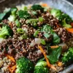 Viral high protein beef bowl - the image is a close-up of a bowl of food. the bowl is made of dark brown ceramic and is placed on a dark wooden table. the food appears to be a stir-fry dish with quinoa, broccoli, carrots, and green onions. the quinoa is a light brown color and is mixed with various vegetables such as sliced carrots, green onions, and spinach. the vegetables are bright green and look fresh and vibrant. the dish looks appetizing and ready to eat.
