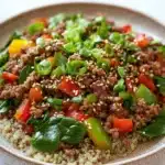 Viral high protein beef bowl - the image is a close-up of a bowl of food. the bowl is made of ceramic and has a light brown color. the food appears to be a salad with a variety of ingredients, including quinoa, red bell peppers, green onions, and sliced avocado. the quinoa is a light beige color and is scattered throughout the dish. the vegetables are bright red and appear to be fresh and vibrant. the dish is garnished with a sprinkle of sesame seeds. the background is a white marble countertop.