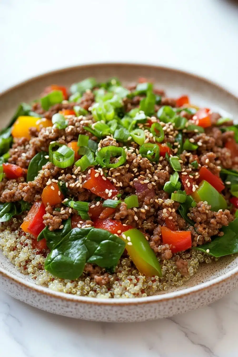 Viral high protein beef bowl - the image is a close-up of a bowl of food. the bowl is made of ceramic and has a light brown color. the food appears to be a salad with a variety of ingredients, including quinoa, red bell peppers, green onions, and sliced avocado. the quinoa is a light beige color and is scattered throughout the dish. the vegetables are bright red and appear to be fresh and vibrant. the dish is garnished with a sprinkle of sesame seeds. the background is a white marble countertop.